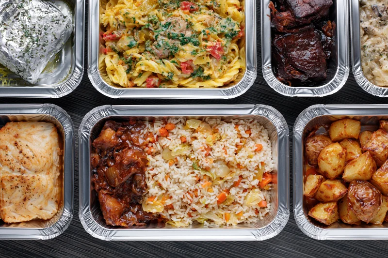 Multiple aluminum foil takeout containers filled with assorted meals arranged on a dark surface