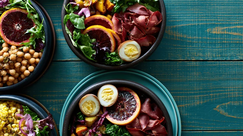 Multiple dark stone bowls filled with salads topped with blood oranges, eggs, bacon, and chickpeas arranged on a teal wooden table