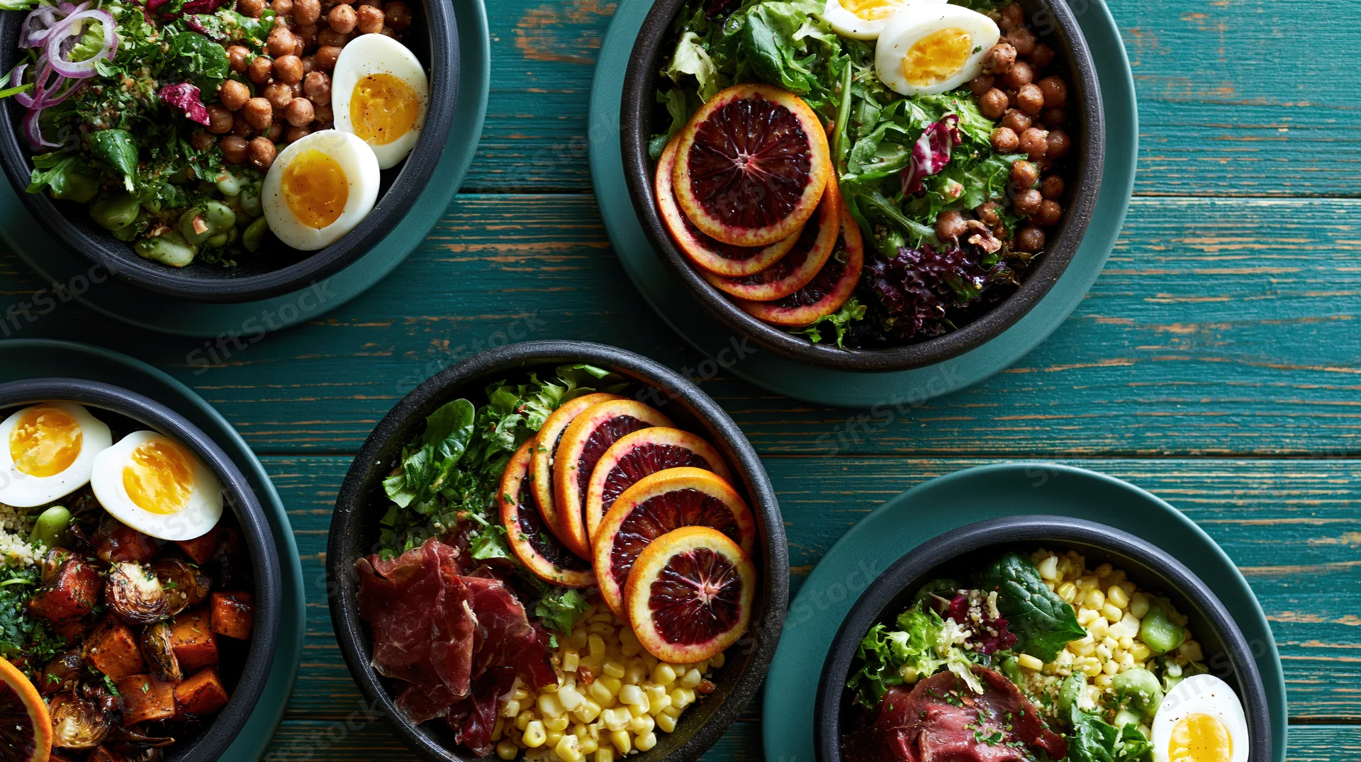 Multiple dark stone bowls filled with colorful salads arranged on a teal wooden table