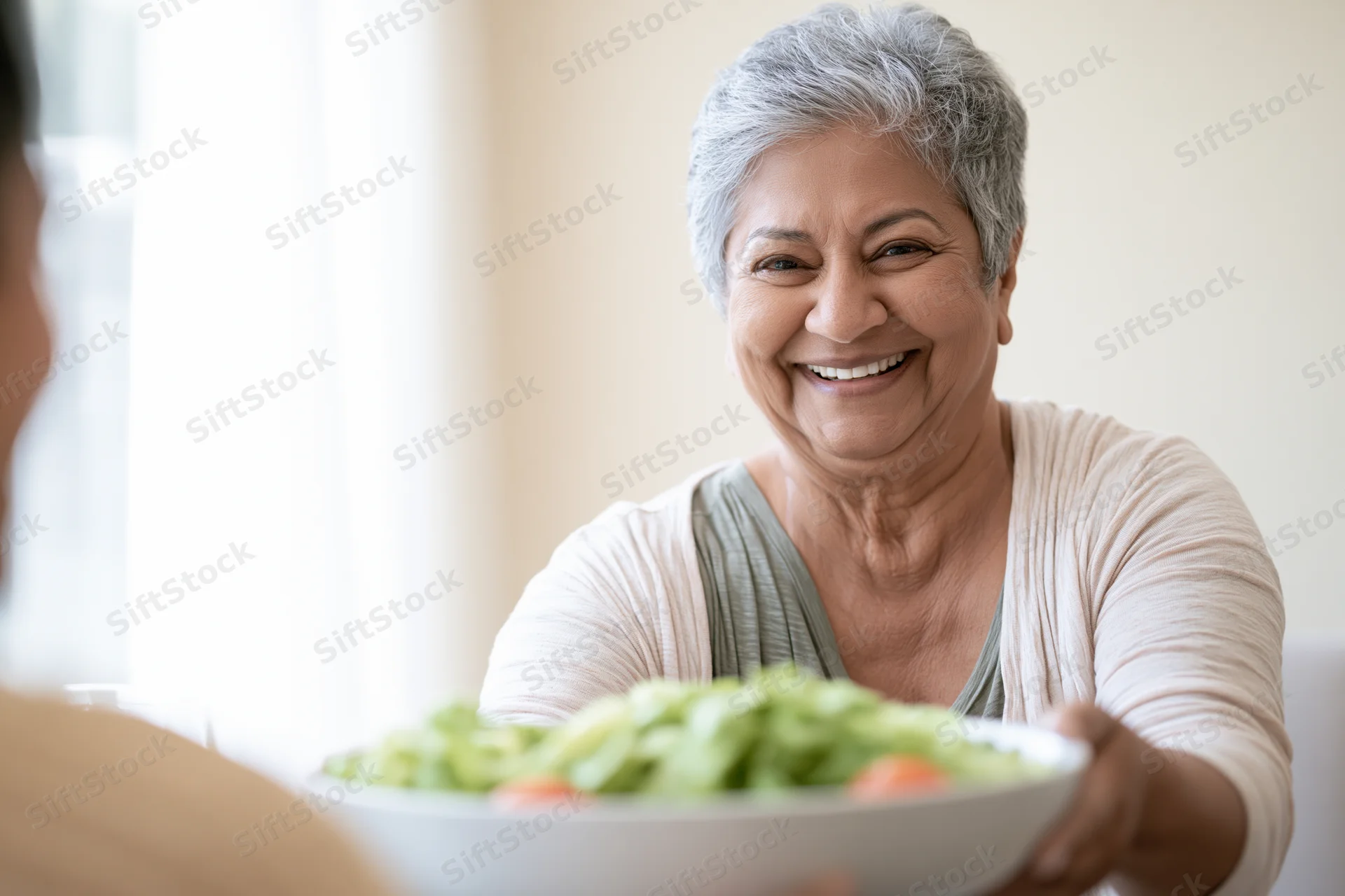 Senior southasian woman smiling while offering a bowl of salad at home