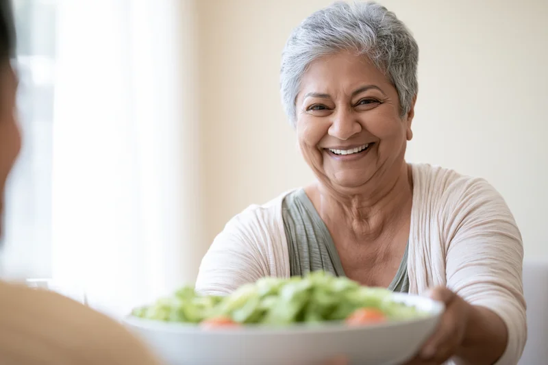 Senior southasian woman smiling while offering a bowl of salad at home
