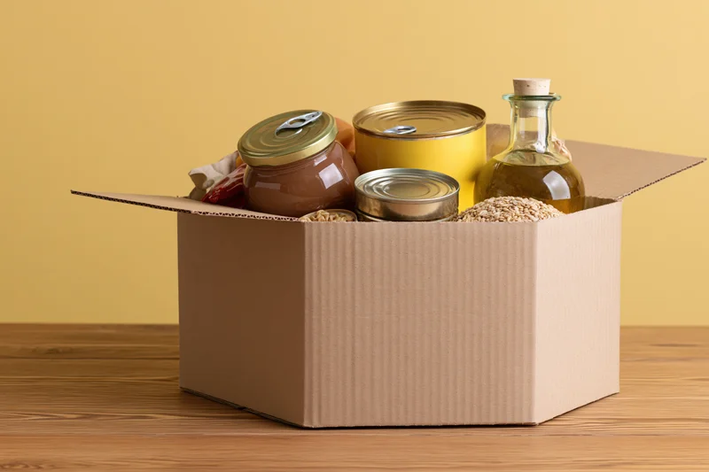 Open cardboard box filled with canned goods and jars on wooden table