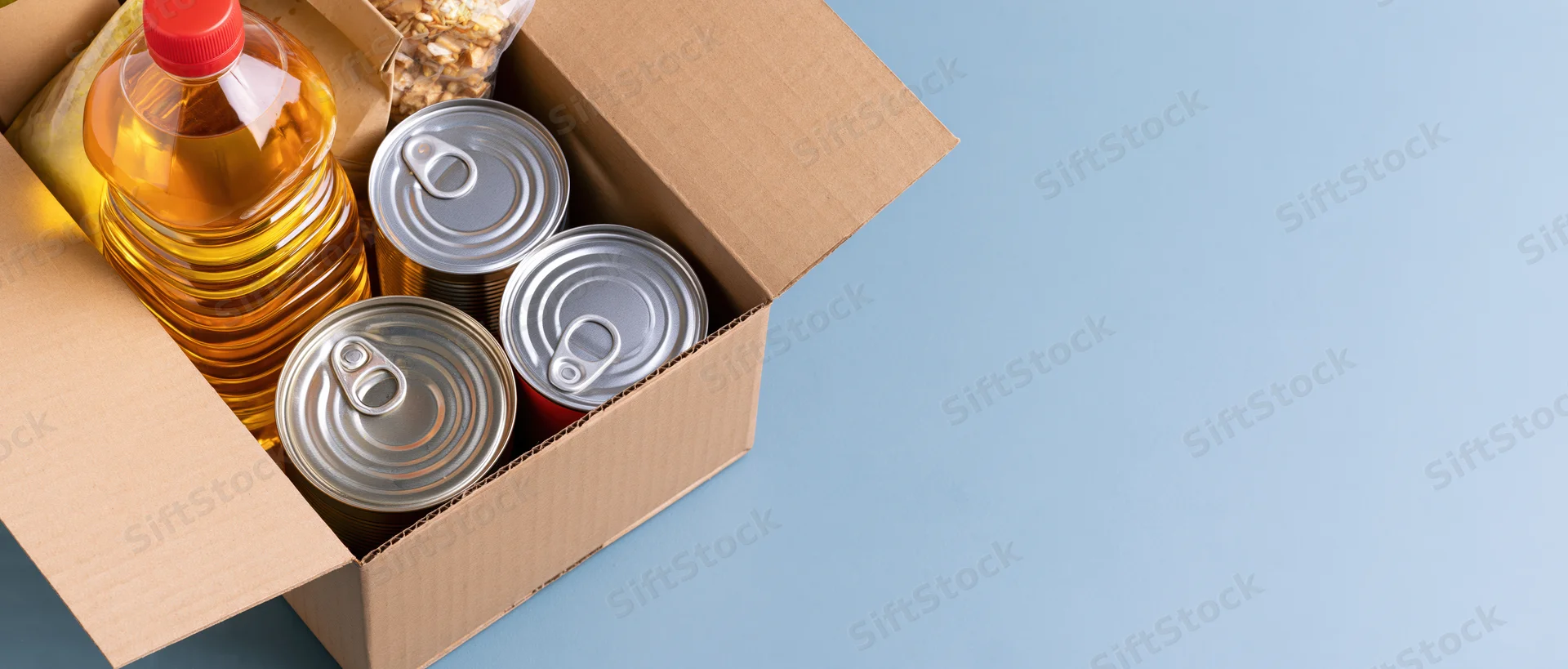 Open cardboard box filled with canned goods and cooking oil on blue surface