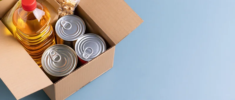 Open cardboard box filled with canned goods and cooking oil on blue surface