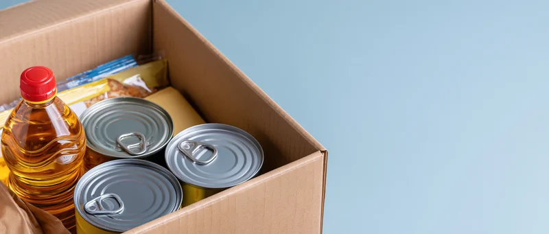 Open cardboard box filled with canned goods and cooking oil against a light blue background