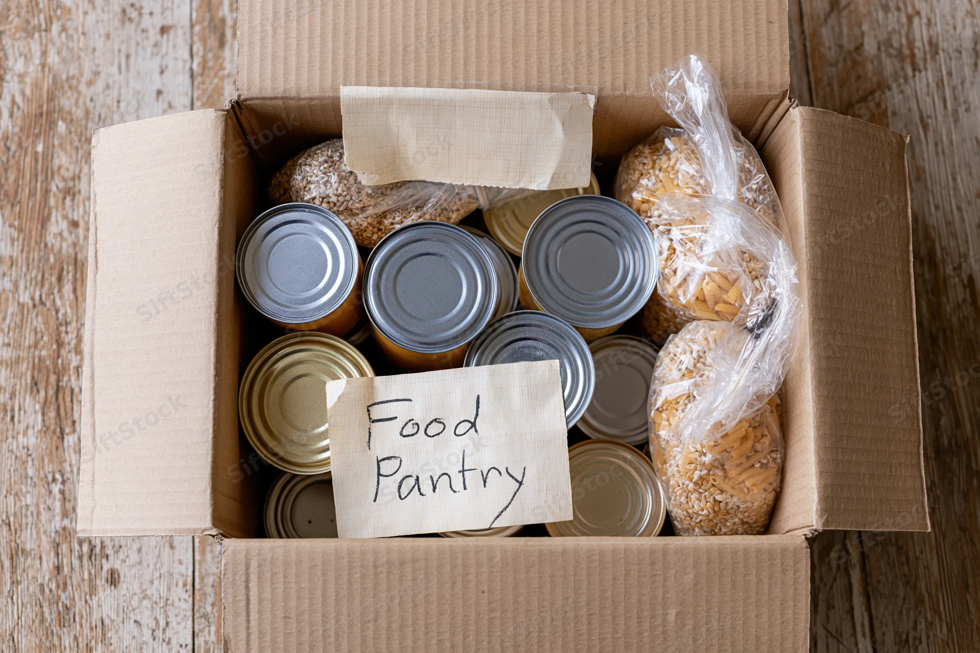 Open cardboard box filled with canned goods and dry food labeled food pantry on wooden surface