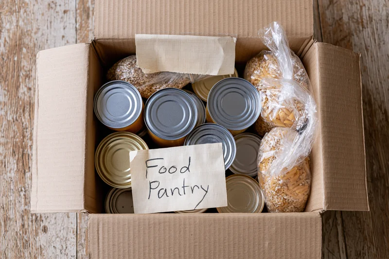 Open cardboard box filled with canned goods and dry food labeled food pantry on wooden surface