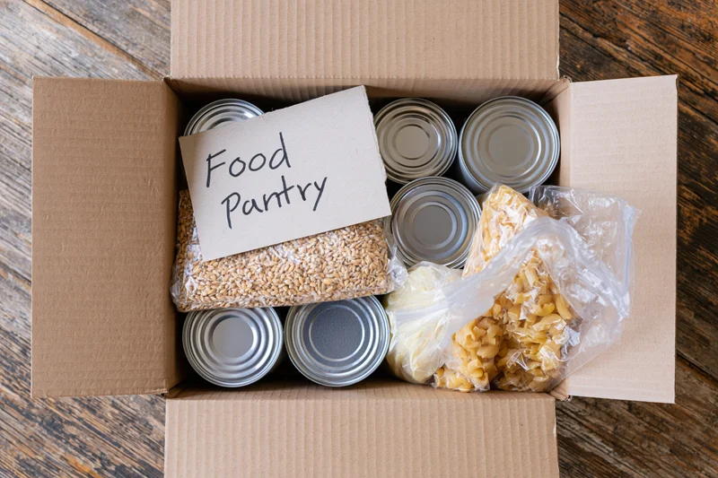 Open cardboard box filled with canned goods and dry food labeled pantry on wooden surface