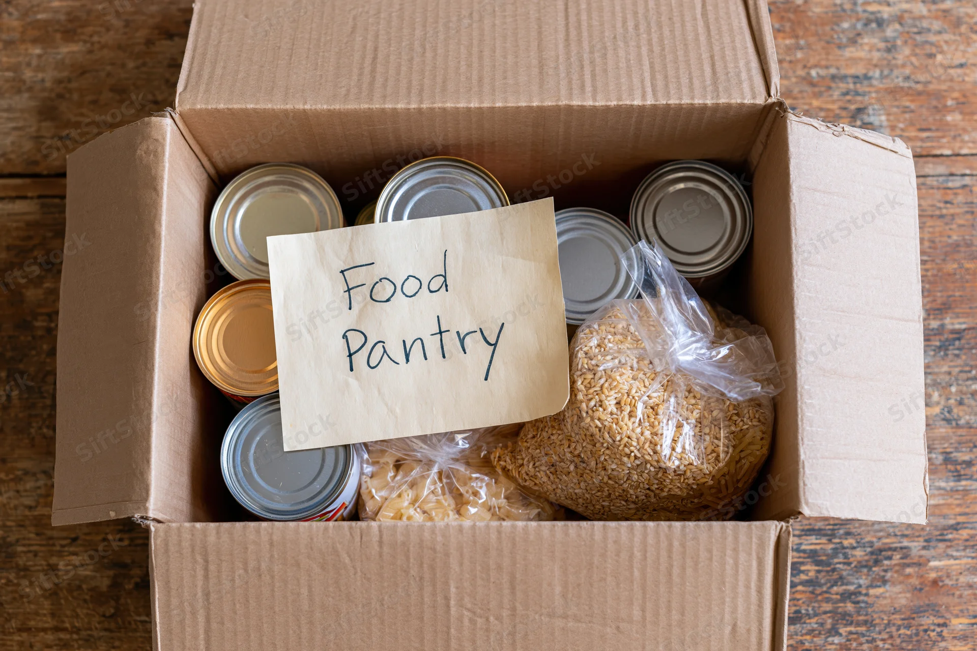 Open cardboard box filled with canned goods and dry food labeled food pantry on wooden surface