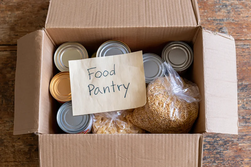 Open cardboard box filled with canned goods and dry food labeled food pantry on wooden surface