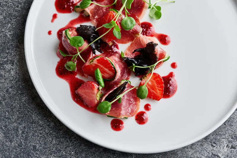 Fine dining appetizer with prosciutto-wrapped strawberries and microgreens on a white plate, garnished with red sauce and herbs, overhead close-up view