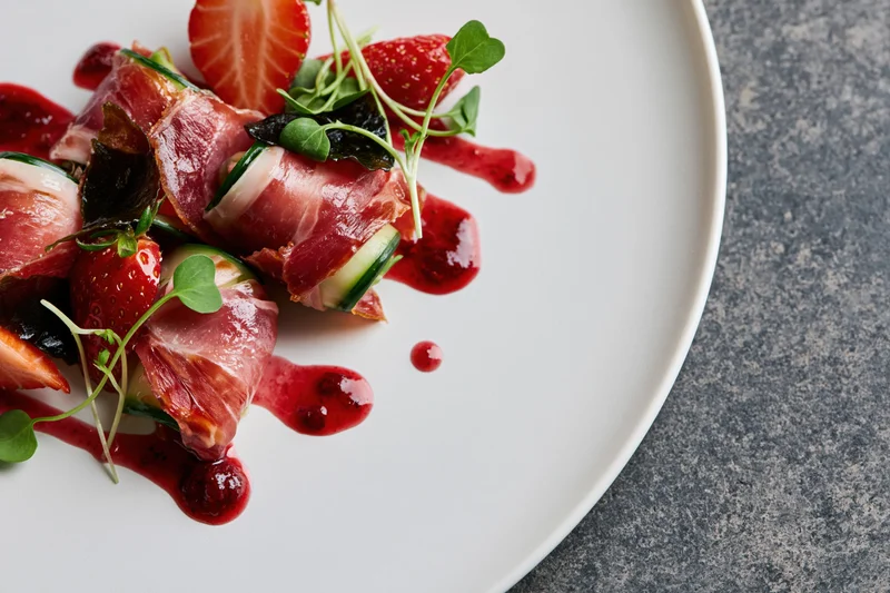 Prosciutto-wrapped appetizer with strawberries and microgreens on a white plate, drizzled with red sauce, overhead close-up on gray stone surface
