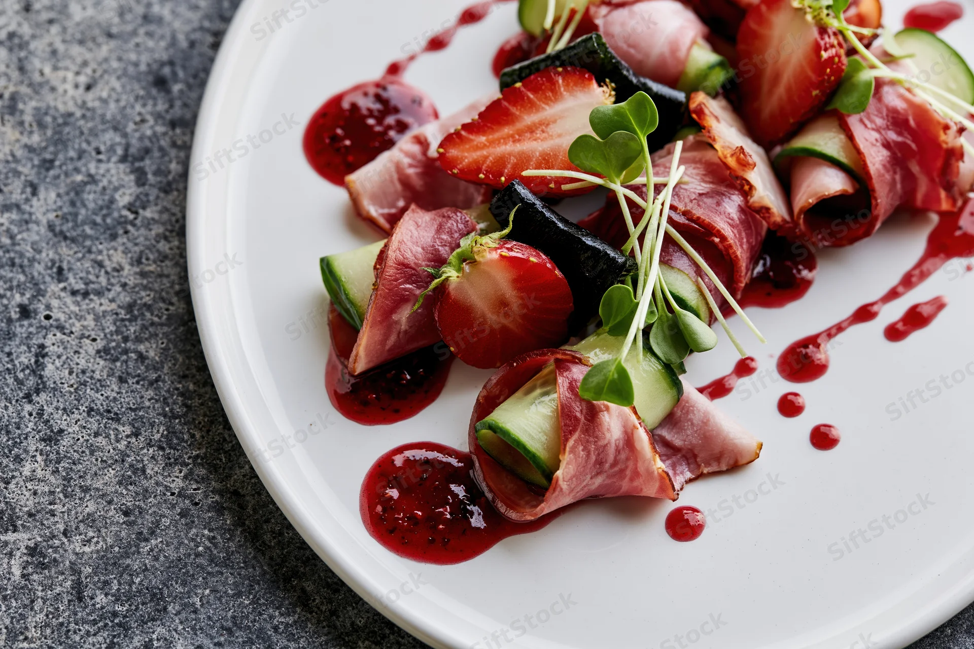 Fine dining appetizer with ham-wrapped cucumber and strawberries on a white plate with red sauce drizzle and microgreens garnish