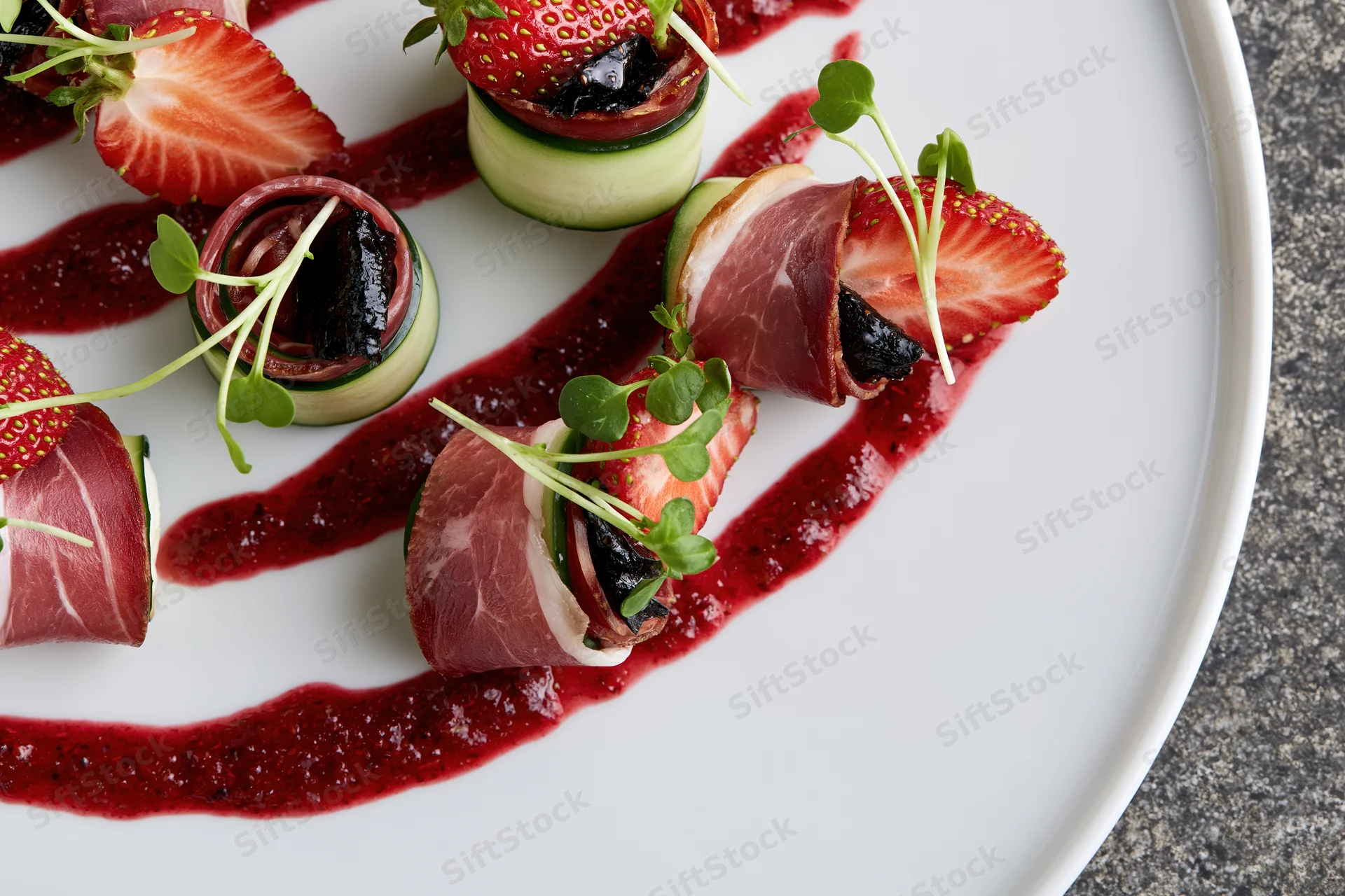 Fine dining appetizer with prosciutto-wrapped strawberries and cucumber rolls on a white plate with red sauce and microgreens garnish