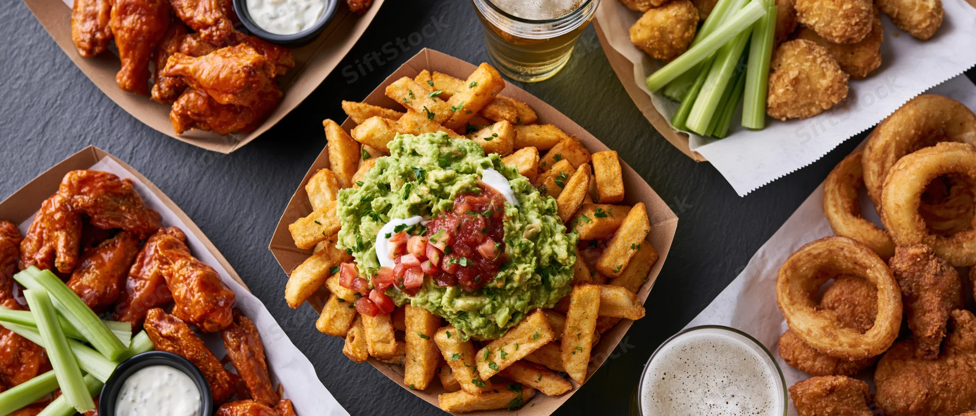 American bar food spread with wings, fries, onion rings and beer on dark slate surface