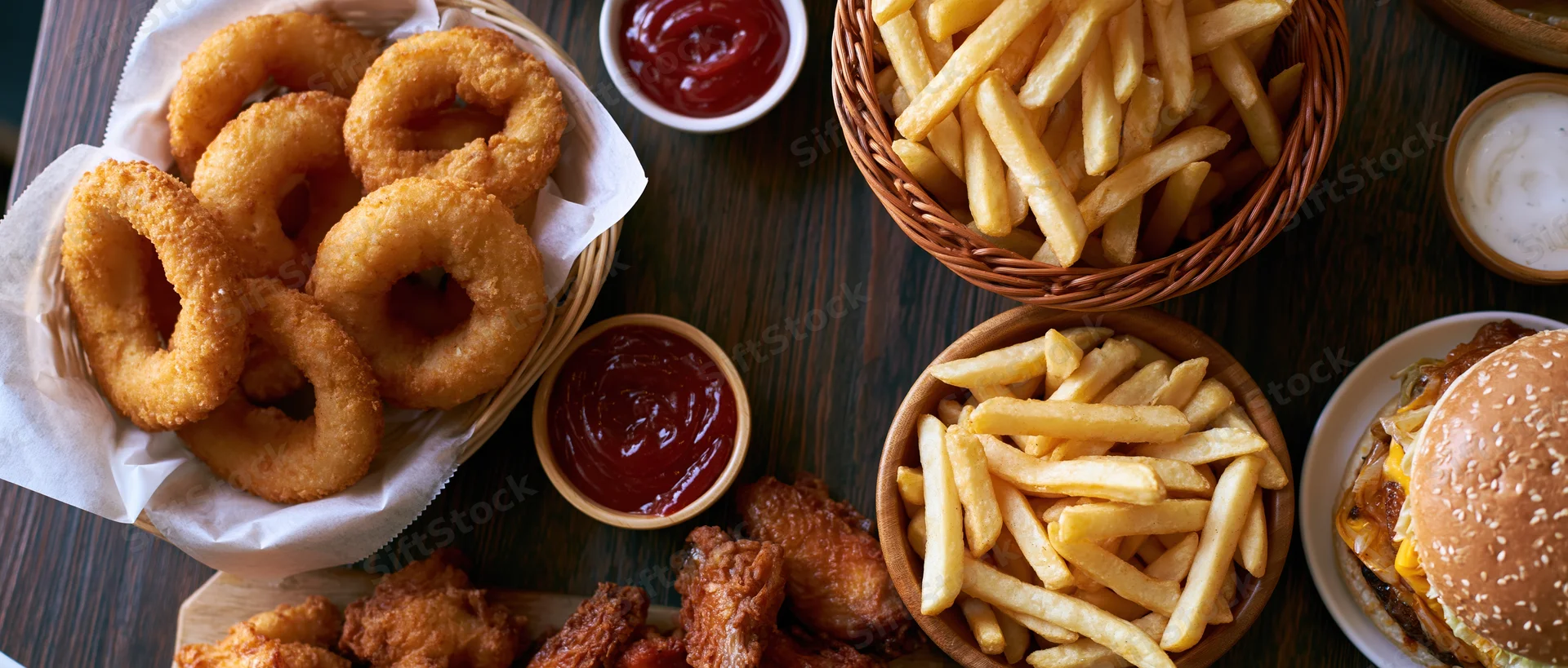 Overhead flat-lay of american fast food spread on dark wooden table featuring burger, fries, onion rings, chicken wings, and condiments