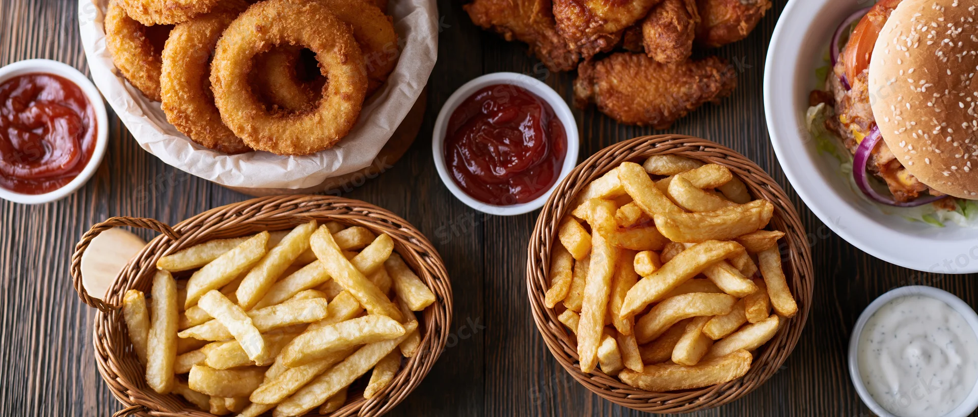 Overhead flat-lay of american fast food including burger, fries, onion rings and chicken wings on dark wooden table