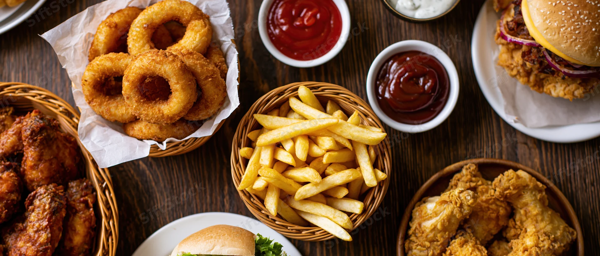 Overhead flat-lay of american fast food spread on dark wooden table featuring burger, fries, onion rings, fried chicken, and condiments