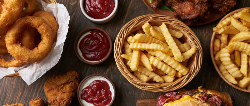 Overhead flat-lay of american fast food including fried chicken, crinkle-cut fries, onion rings, and ketchup on a dark wooden table