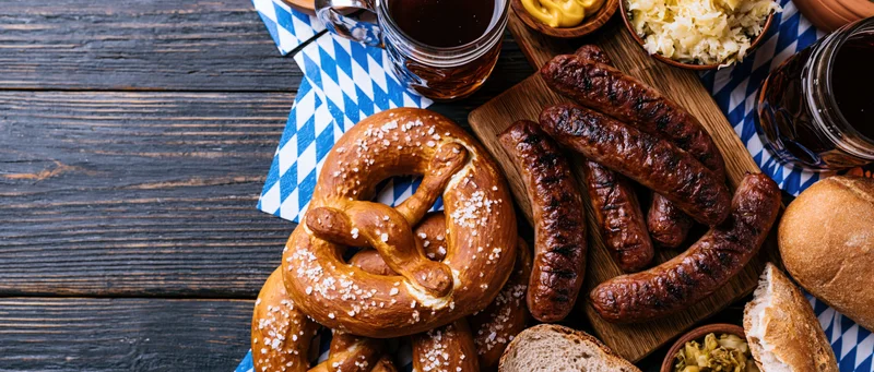 Overhead view of oktoberfest food spread with sausages, pretzels and beer on a dark wooden table with blue and white checkered napkin