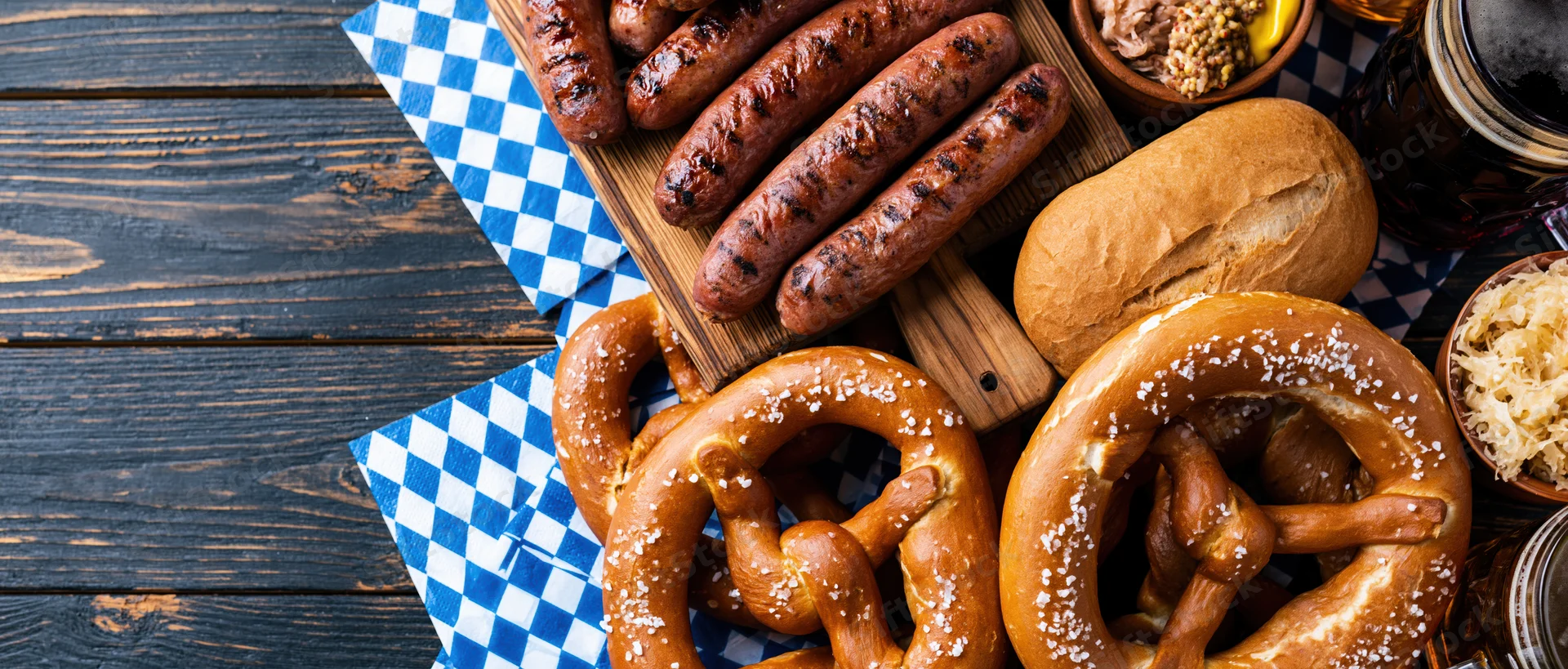 Grilled sausages and pretzels with bread and condiments arranged on a dark wooden table with blue checkered napkins for an Oktoberfest food spread