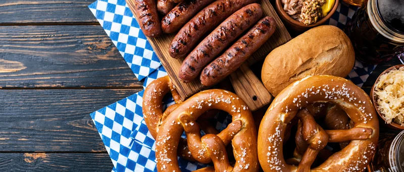 Grilled sausages and pretzels with bread and condiments arranged on a dark wooden table with blue checkered napkins for an Oktoberfest food spread