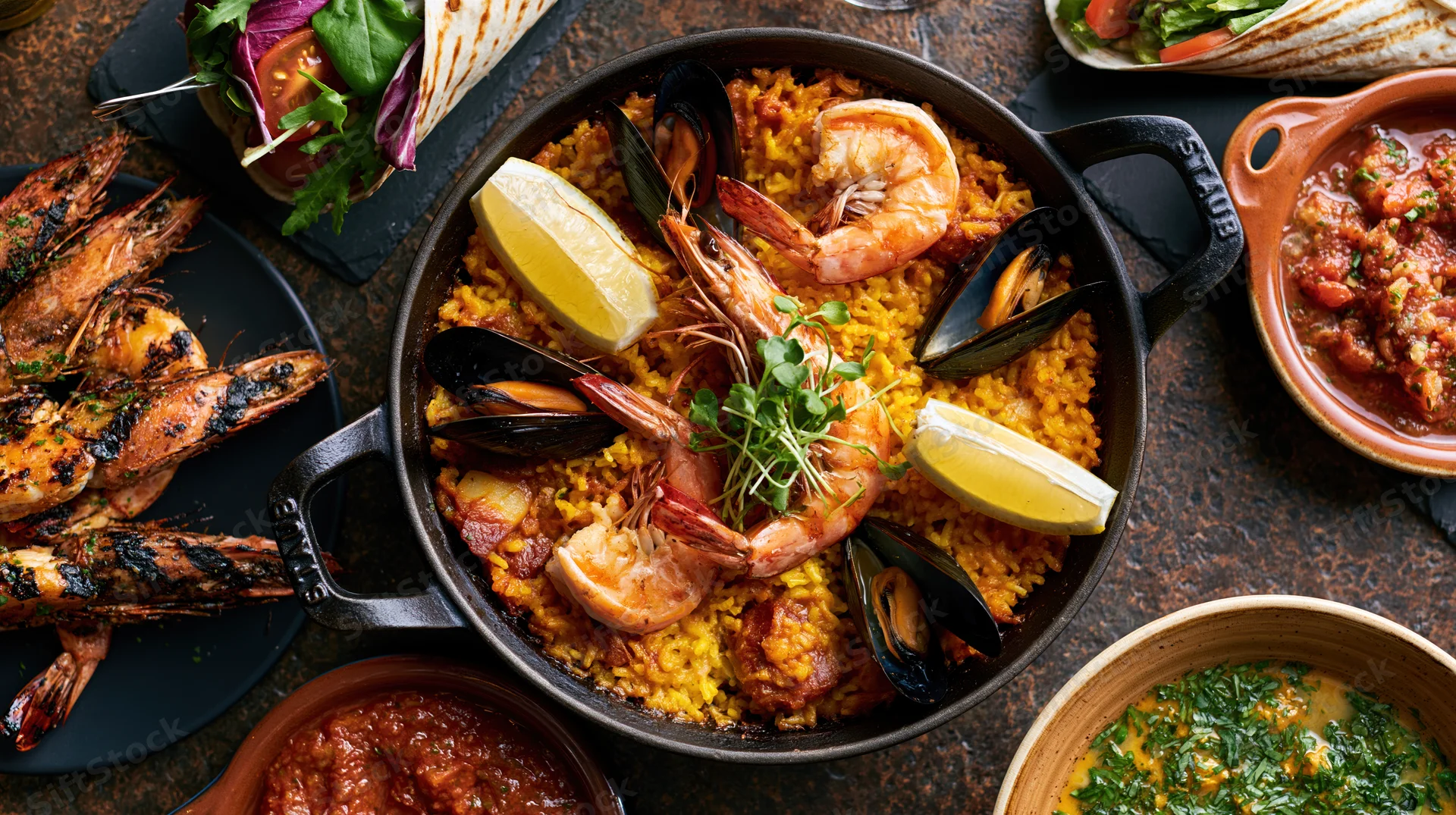 Spanish seafood paella in a cast iron skillet with shrimp and mussels, surrounded by side dishes on a dark stone table