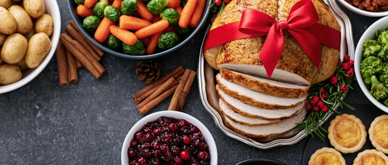 Sliced roasted turkey with red bow and holiday side dishes arranged on dark table in overhead flat lay
