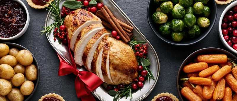 Sliced roasted turkey garnished with rosemary and cranberries on a silver platter surrounded by holiday side dishes on a dark slate table