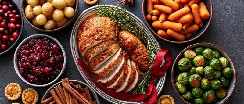 Sliced roasted turkey garnished with herbs and red ribbon on a dark surface surrounded by holiday side dishes including cranberry sauce, carrots, brussels sprouts, potatoes, and cinnamon sticks