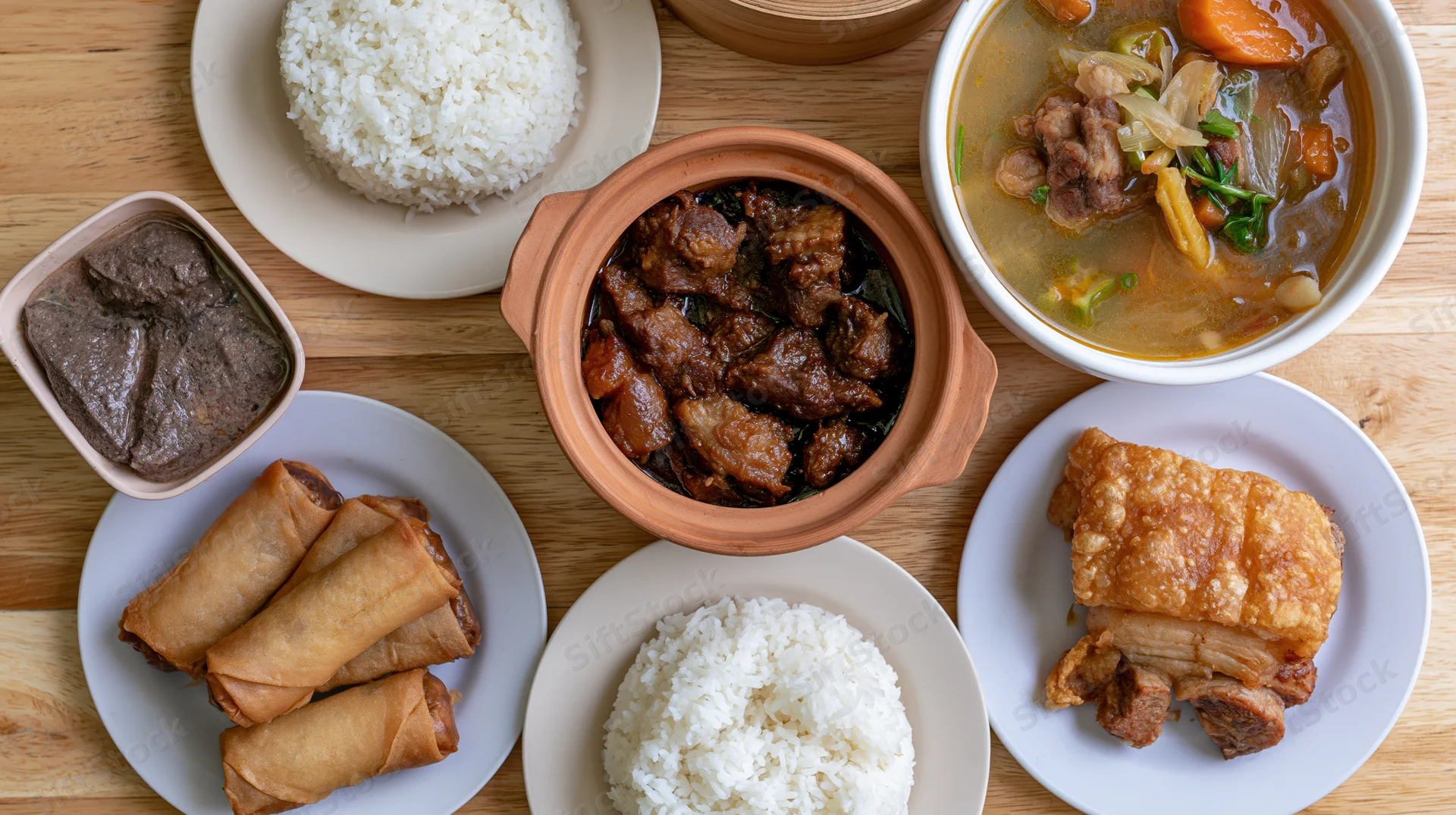 Overhead flat-lay of a Filipino home-cooked meal spread on a wooden table with rice, adobo, sinigang, lumpia, and lechon on plates and bowls