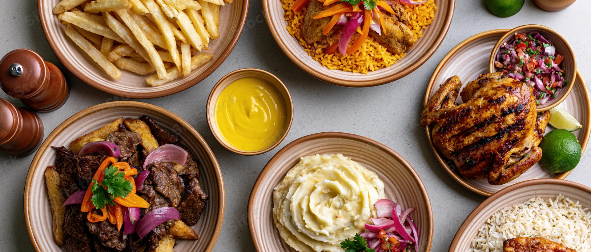 Overhead flat-lay of a peruvian restaurant spread with multiple dishes including beef, chicken, fries, rice, mashed potato, sauce, and garnishes on a table