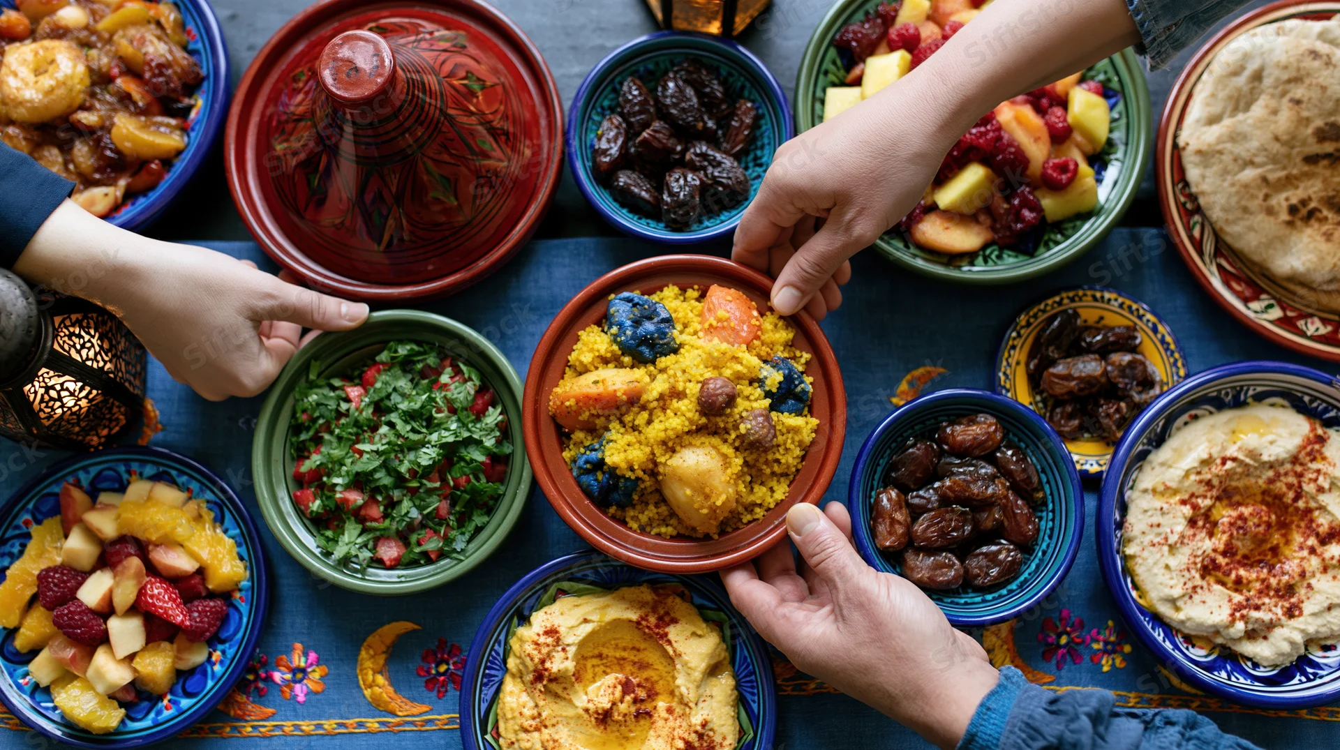 Hands sharing dishes of couscous, dates, hummus and salad on a patterned table during iftar dinner