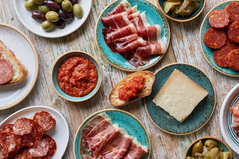 Overhead flat-lay of a spanish tapas spread on a weathered wooden table with plates of ham, cheese, olives, salami, and tomato sauce