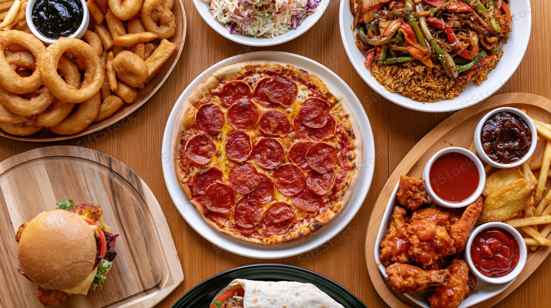 Overhead view of a large spread of casual restaurant food including pizza, burger, chicken wings, fries, onion rings, and side dishes on a wooden table