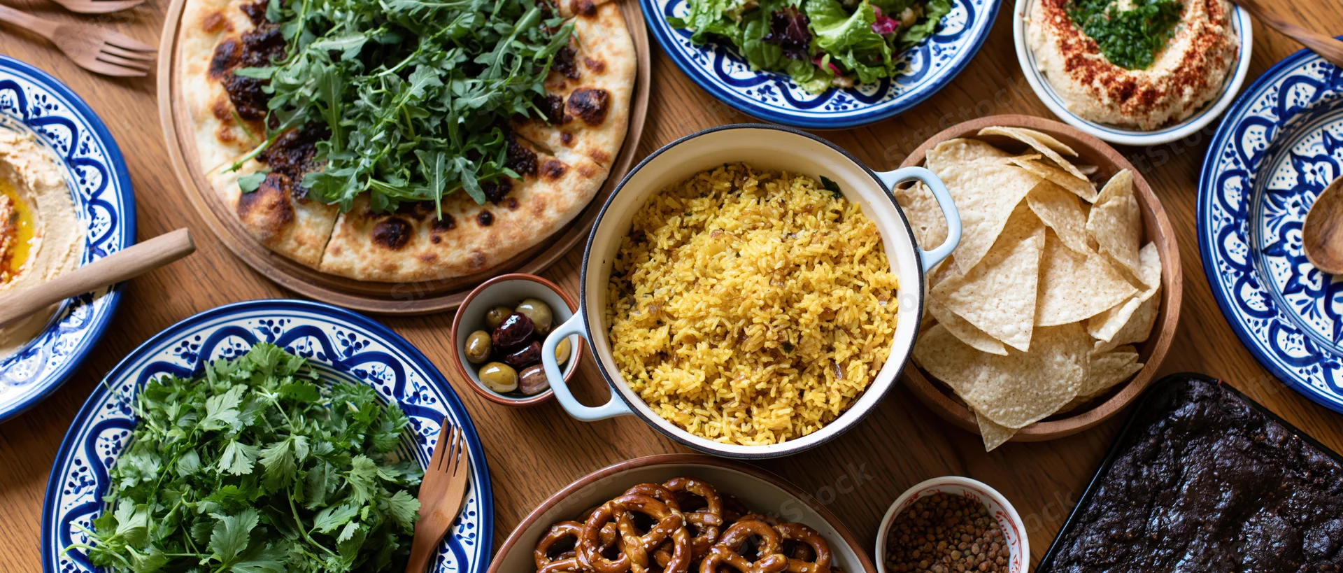 Overhead view of a wooden dining table set with various dishes including pizza, hummus, rice, chips, and salads arranged for a shared meal