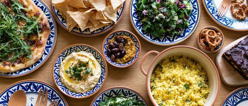 Overhead flat-lay of a wooden dining table with various dishes including hummus, salad, rice, olives, chips, and dessert arranged on ceramic plates and bowls