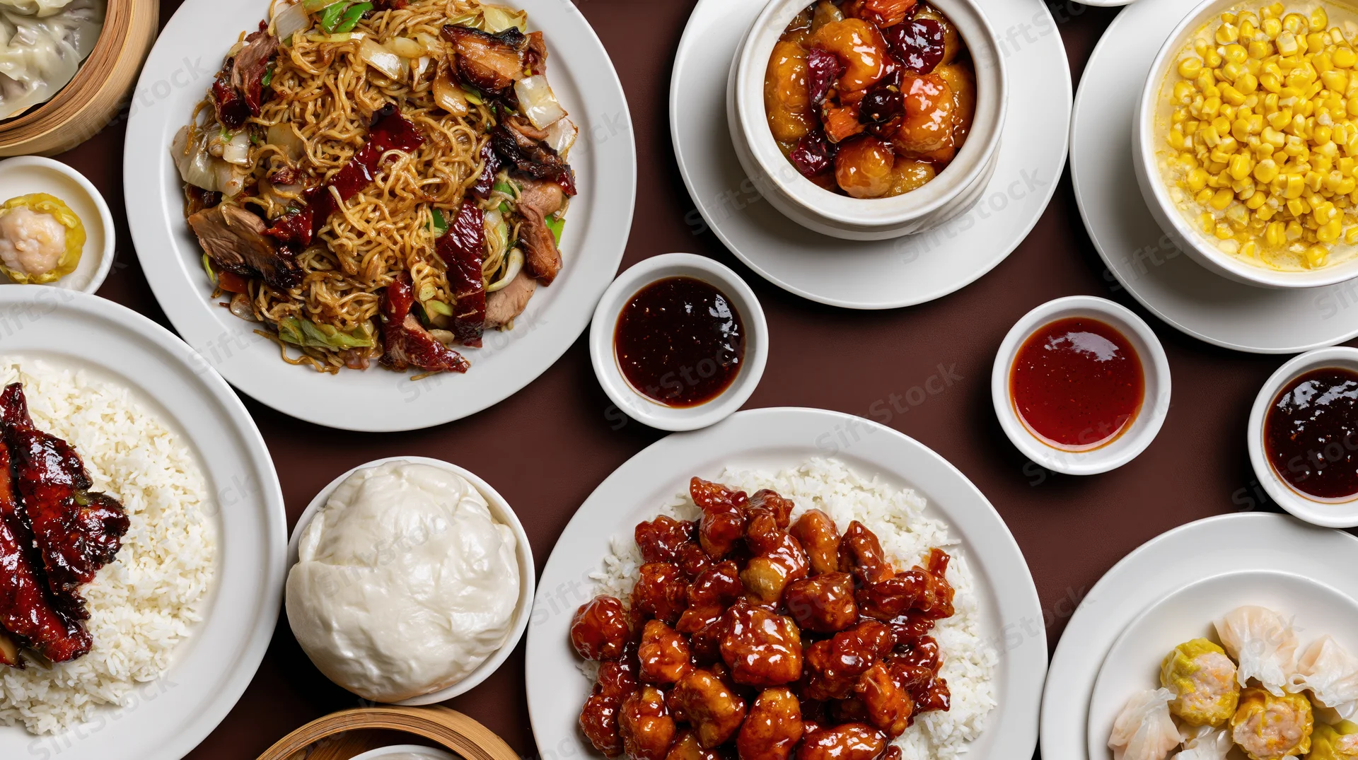 Overhead flat-lay of various chinese restaurant dishes arranged on a brown table including noodles, rice, sweet and sour chicken, dumplings, corn, and sauces