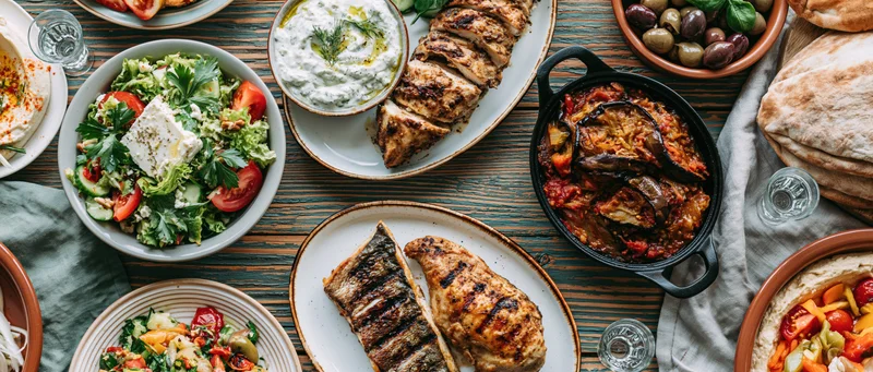 Overhead view of greek mezze dishes arranged on a wooden table with grilled chicken, fish, salad, tzatziki, and pita bread