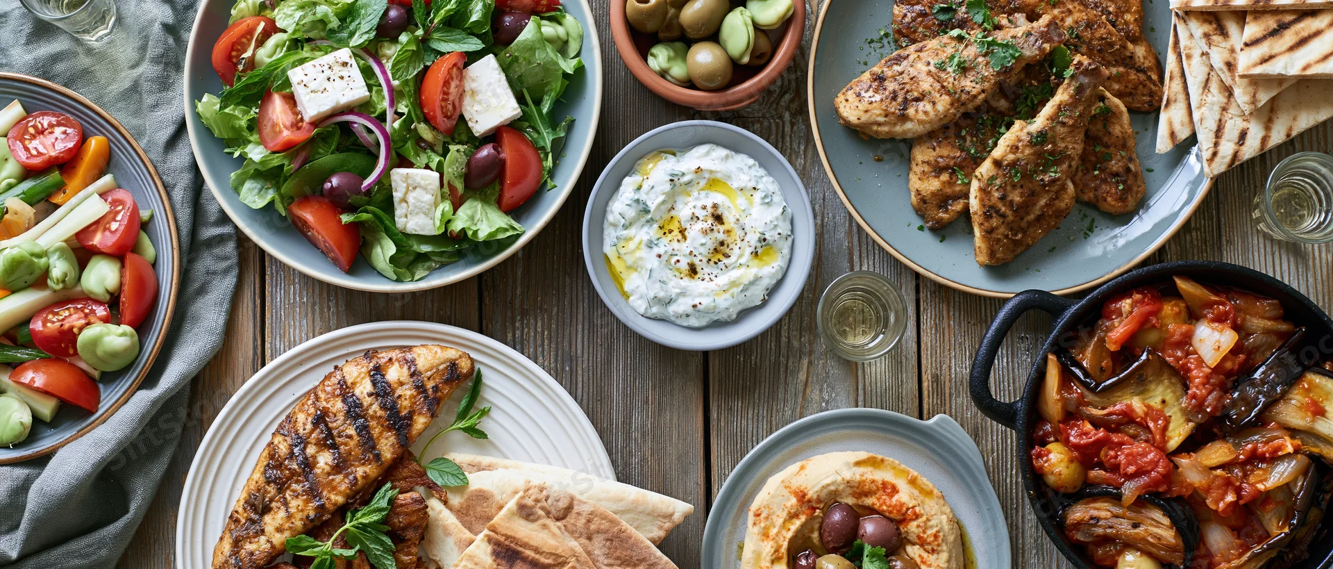 Overhead view of greek mezze dishes arranged on a rustic wooden table with grilled chicken, salad, tzatziki, hummus, pita, olives, and vegetables