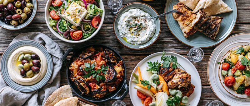 Overhead view of greek mezze dishes arranged on a rustic wooden table with hummus, tzatziki, grilled chicken, eggplant, olives, and pita bread
