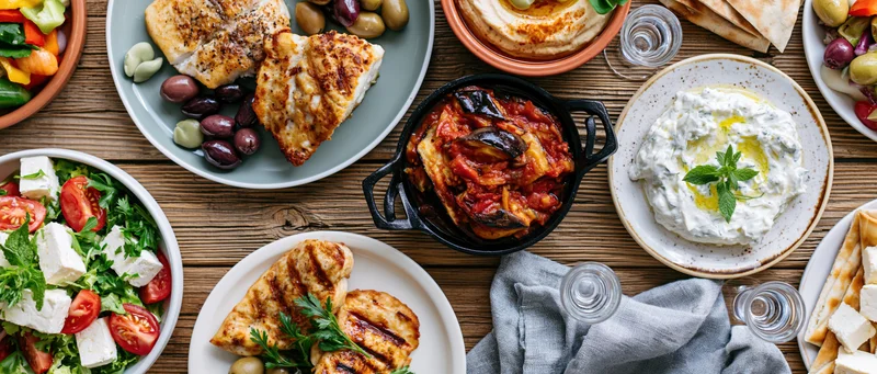 Overhead view of greek mezze dishes arranged on a rustic wooden table with plates of salad, grilled fish, hummus, tzatziki, and pita bread