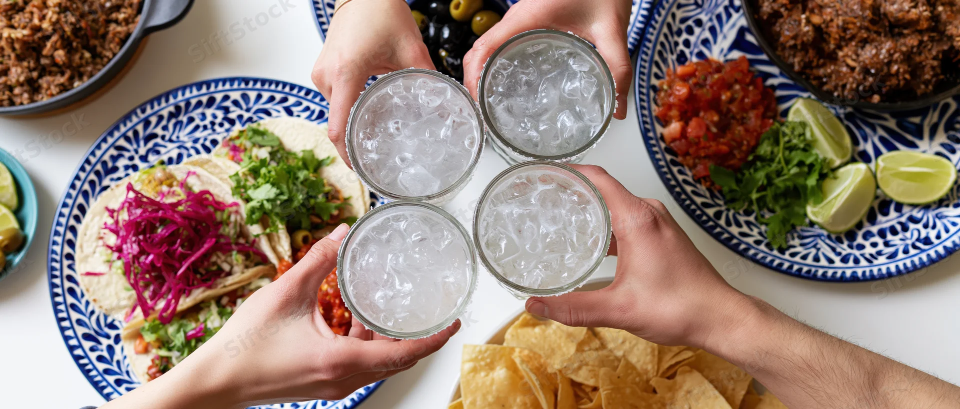 Four hands toasting with glasses of ice water over a taco spread on a white table with blue patterned plates