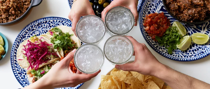 Four hands toasting with glasses of ice water over a taco spread on a white table with blue patterned plates
