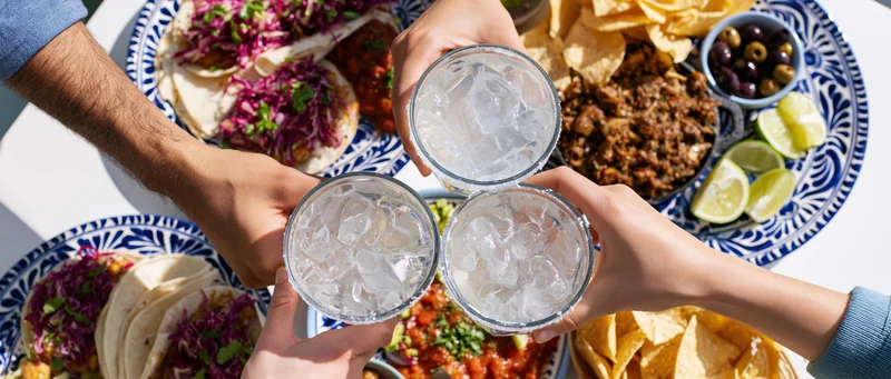 Hands toasting with iced drinks over a taco spread on a white table