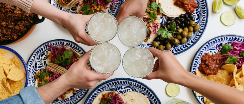 Four hands toasting margaritas over a taco spread on a white table with blue patterned plates