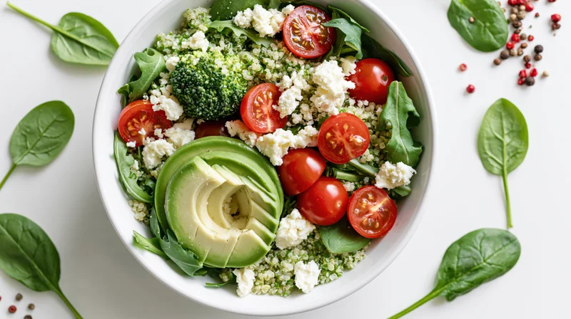Overhead view of a white bowl filled with avocado, cherry tomatoes, spinach, quinoa and feta cheese on a white surface