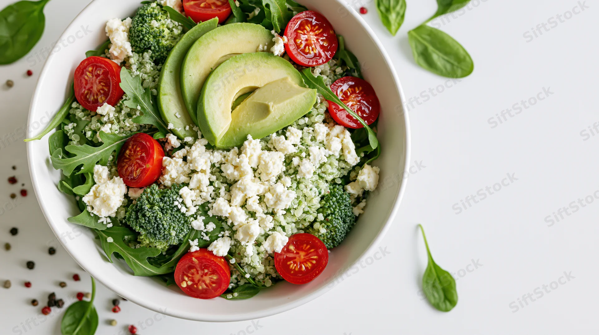 Overhead view of a white ceramic bowl filled with avocado, cherry tomato, broccoli, quinoa, feta, and greens on a white surface