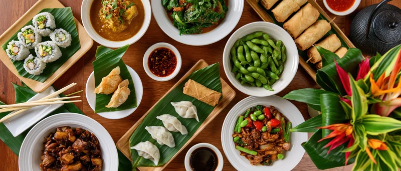 Overhead flat-lay of an asian restaurant spread with sushi, spring rolls, edamame, dumplings, stir fry, curry, sauces, chopsticks, and tropical flowers on wood