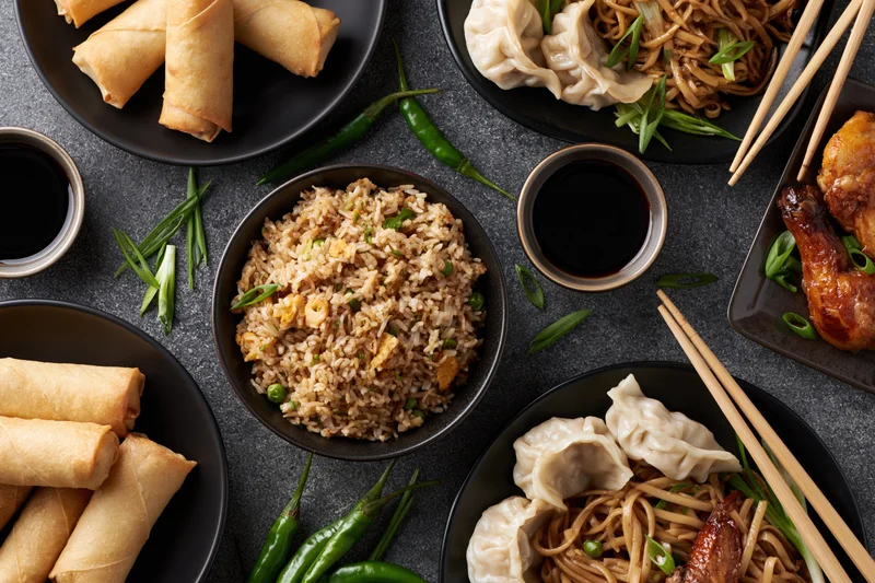Overhead flat-lay of assorted chinese dishes including fried rice, spring rolls, dumplings, noodles, and soy sauce on dark surface